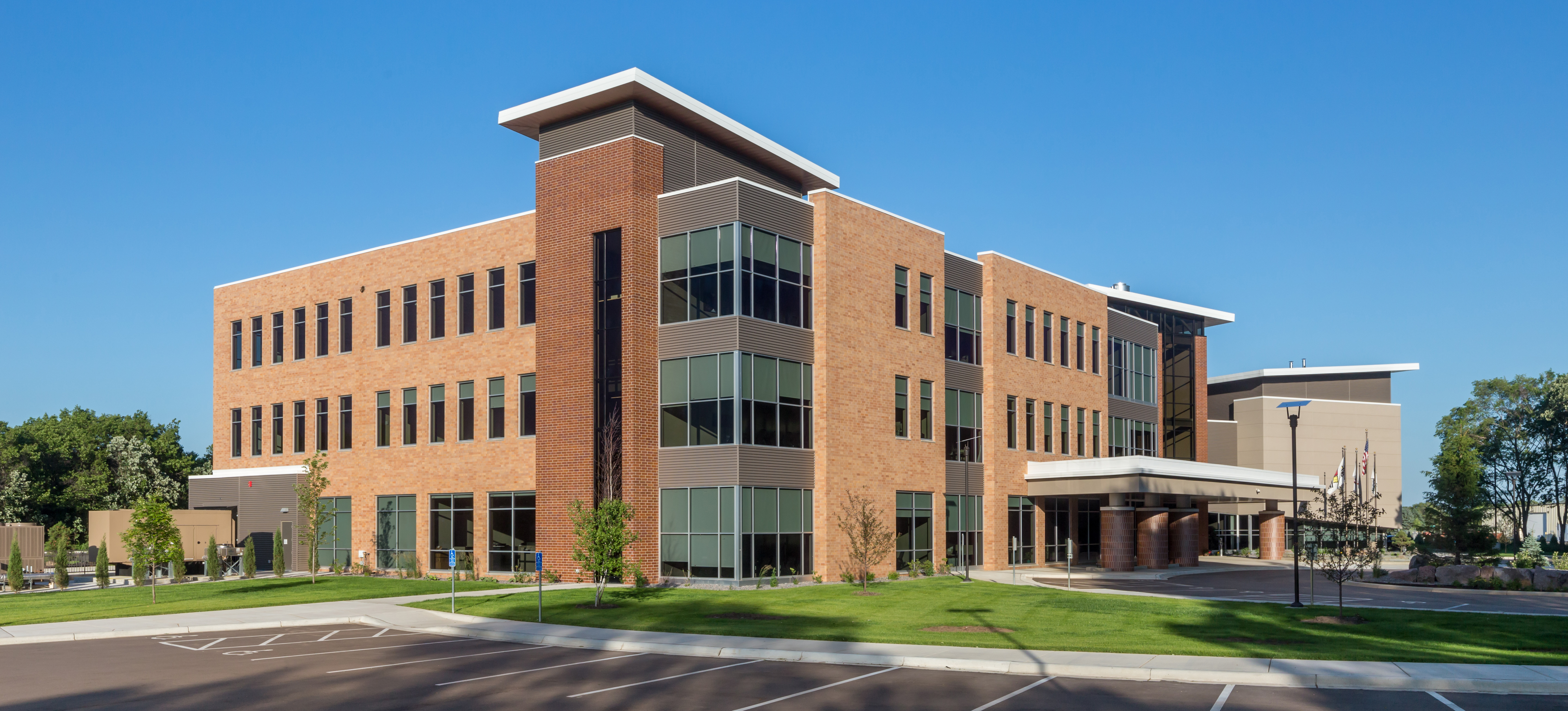 Exterior of a modern three-story brick office building with large windows, landscaped grounds, and a covered entrance under a clear blue sky.