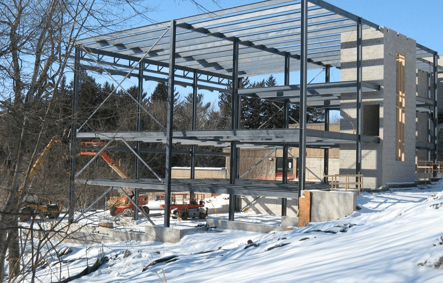 Multi-story steel frame building under construction on a snow-covered site with construction lifts and concrete foundation.