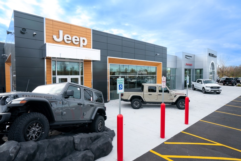 Exterior of a modern Jeep dealership with a gray and wood-accent facade, large glass storefronts, and multiple Jeep vehicles displayed outside the entrance.