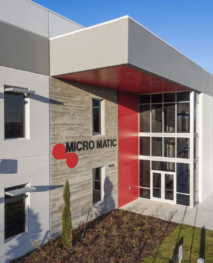 Modern commercial office building exterior with Micro Matic logo, featuring glass entry, red accent panels, and metal wall cladding.