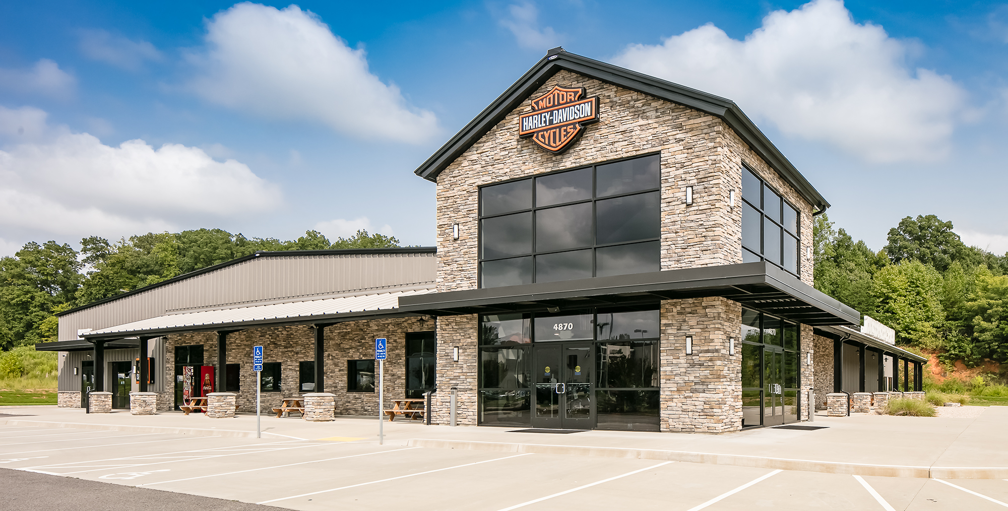 Exterior of a Harley-Davidson motorcycle dealership featuring a stone façade, large glass windows, covered entry, and metal building structure.