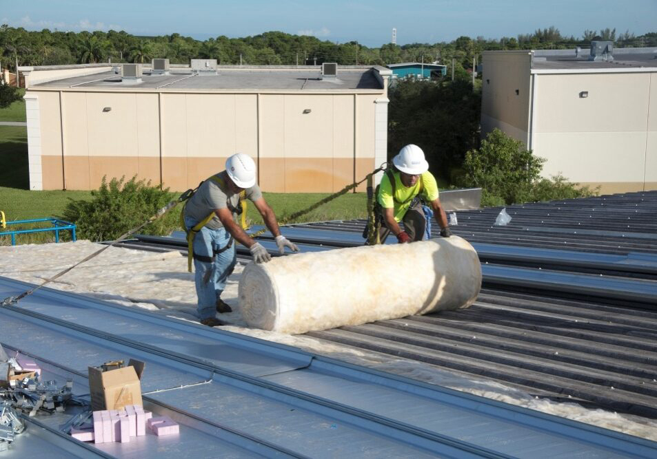 Two workers rolling out fiberglass insulation on a metal roof as part of a commercial reroofing installation.