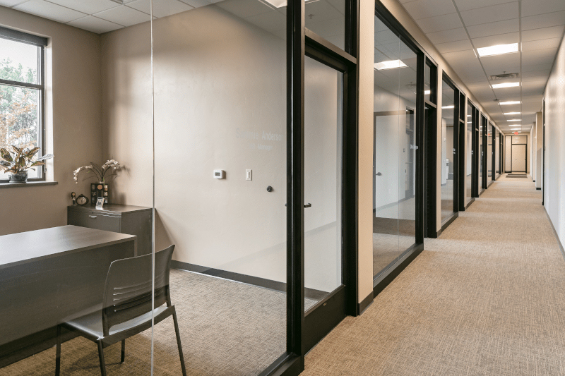 Modern office corridor with glass-walled offices, carpeted flooring, and neutral interior finishes.