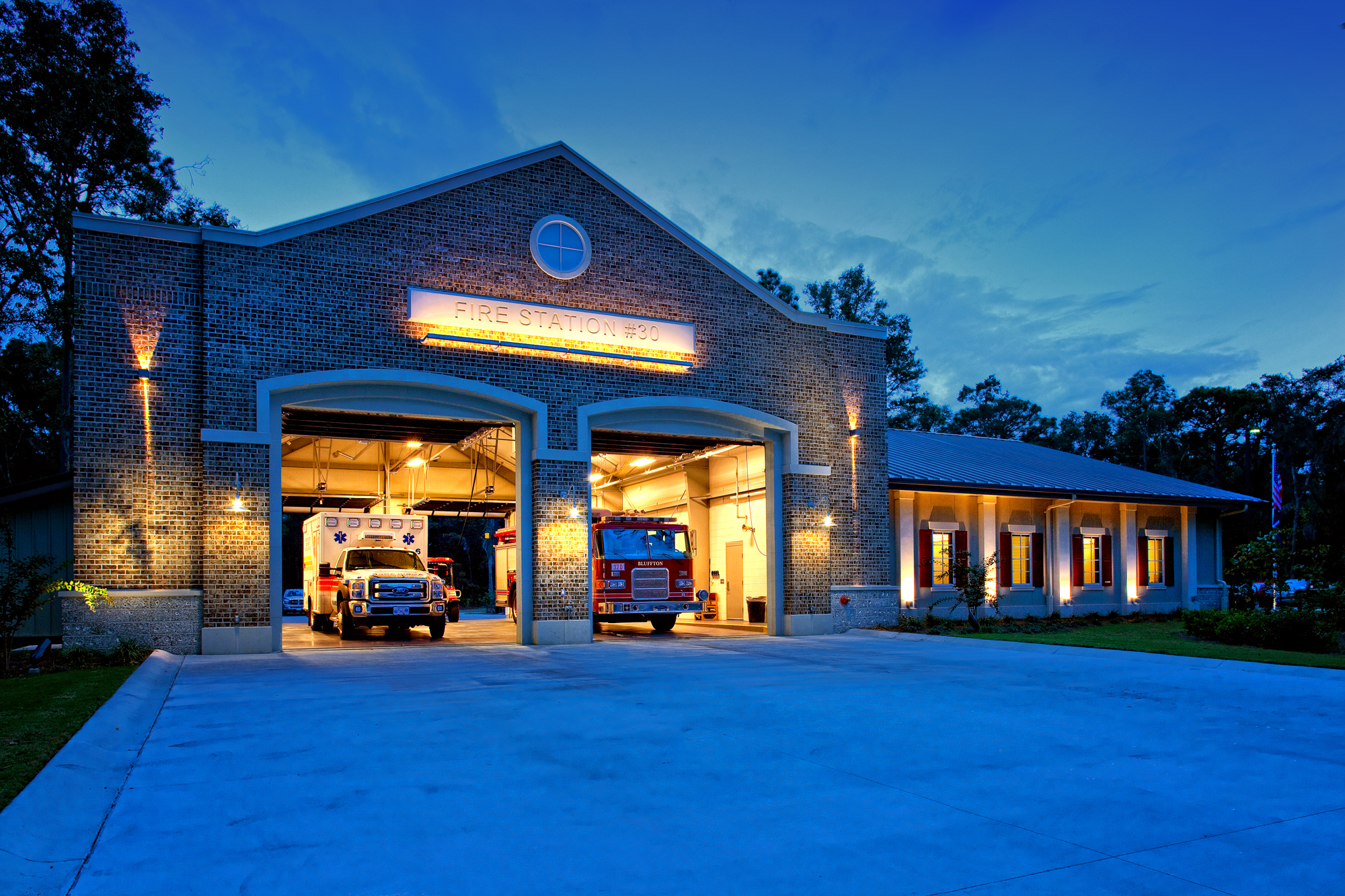 Brick fire station with open garage bays, warm yellow lighting and red emergency vehicles beneath a deep blue evening sky.