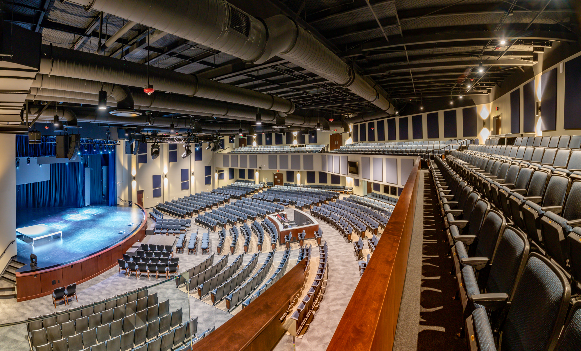 Large auditorium with tiered gray seating, curved wood railings, a blue-lit stage and exposed overhead ductwork.