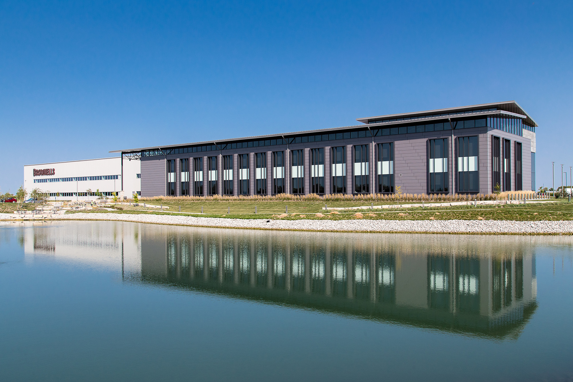 Modern commercial office building with glass façade reflected in a pond under a clear blue sky.