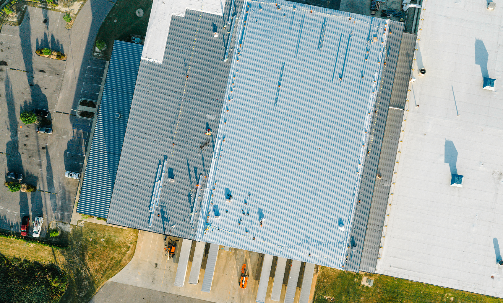 Top-down aerial view of a light gray metal roof with rooftop equipment, loading docks and surrounding parking and landscaping.