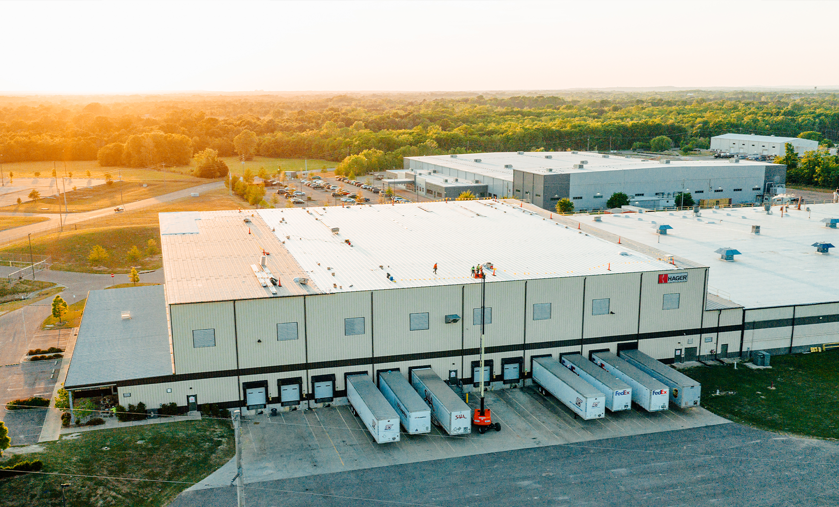 Aerial view of a large white warehouse with a bright white roof, white loading dock trailers and a gray parking area at golden sunset.