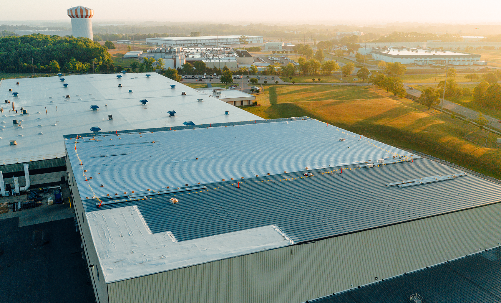Aerial view of a large gray and white industrial building with a light gray roof, surrounded by green fields and commercial buildings at golden sunset.