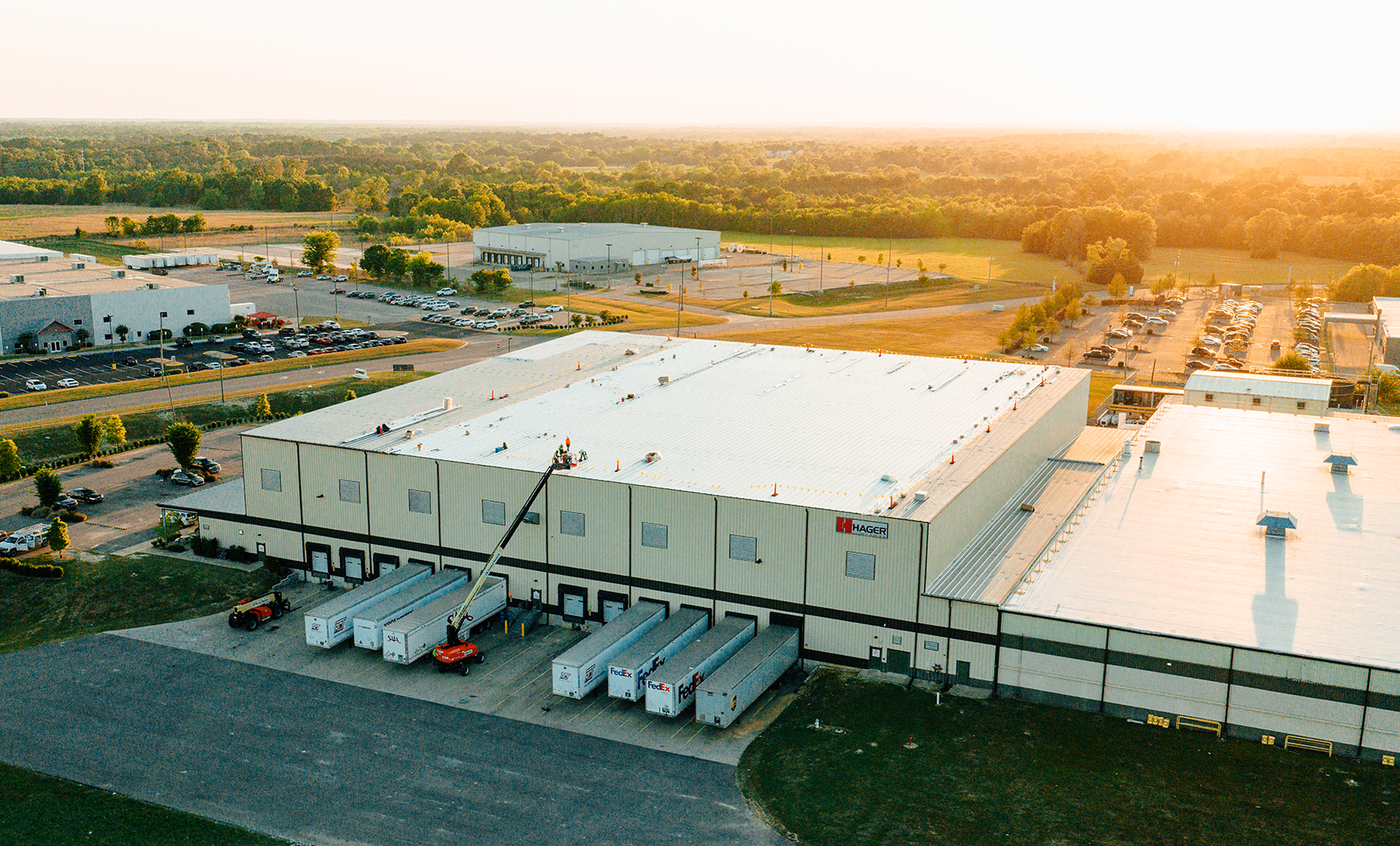 Aerial view of a large white warehouse with a bright white roof, white loading dock trailers and surrounding green landscaping at golden sunset.