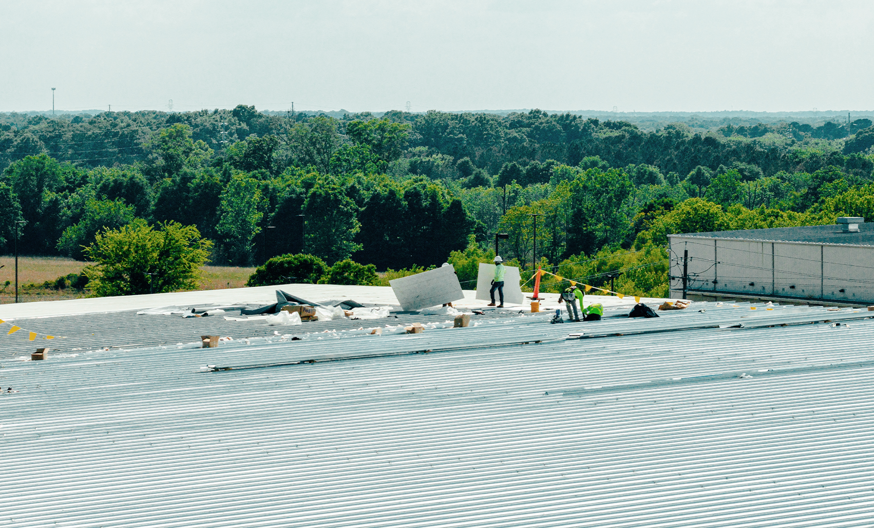 Workers installing a light gray metal roof on a large industrial building with green trees and a clear blue sky in the background.