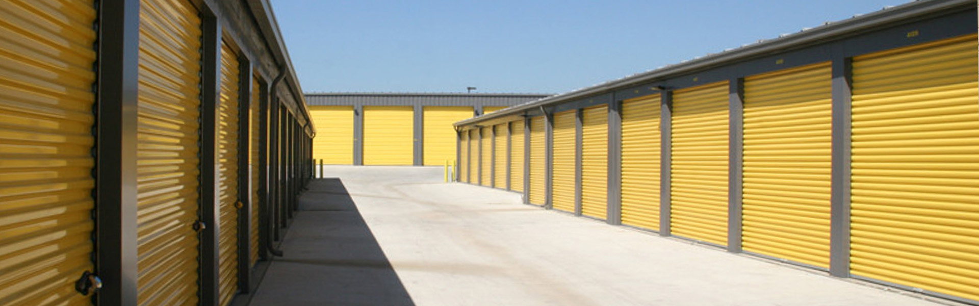 Drive aisle between self-storage buildings with yellow roll-up ribbed doors and gray vertical-rib steel siding
